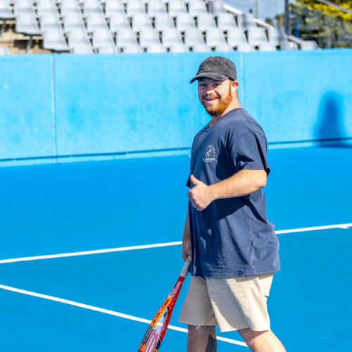 The image shows a person standing on a tennis court, facing the camera. They are smiling and giving a thumbs-up gesture.