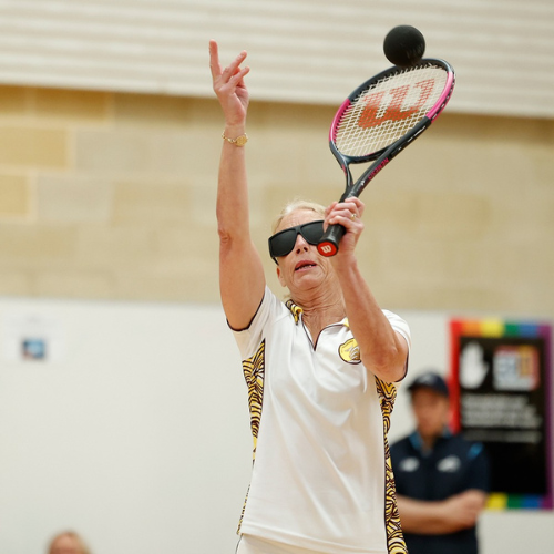 In the image, there is a person wearing a white shirt and black glasses, holding a tennis racket. They are positioned as if they are about to strike a black ball, which is also visible in the image. The ball is used for blind tennis, distinguished by its black color. The person appears focused and ready to engage in the game.