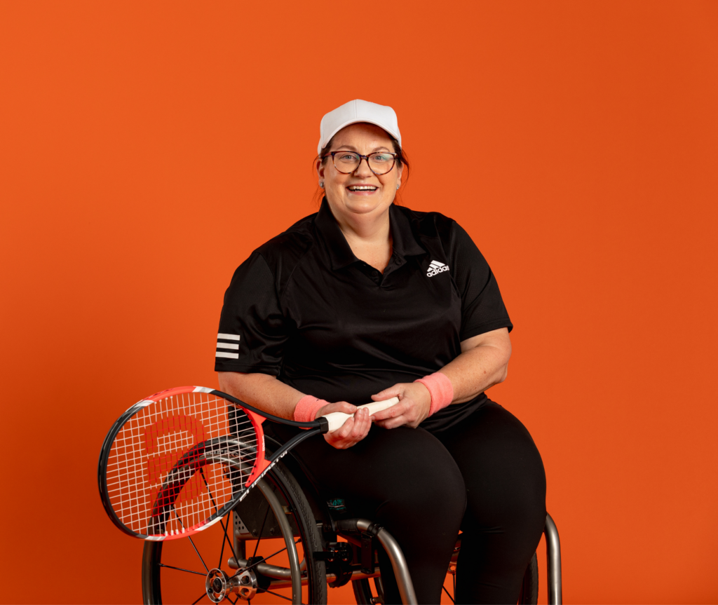 Against a bright orange background, a Caucasian woman with dark hair, wearing a white hat, is sitting in a wheelchair, looking at the camera and smiling. She is holding a tennis racket.