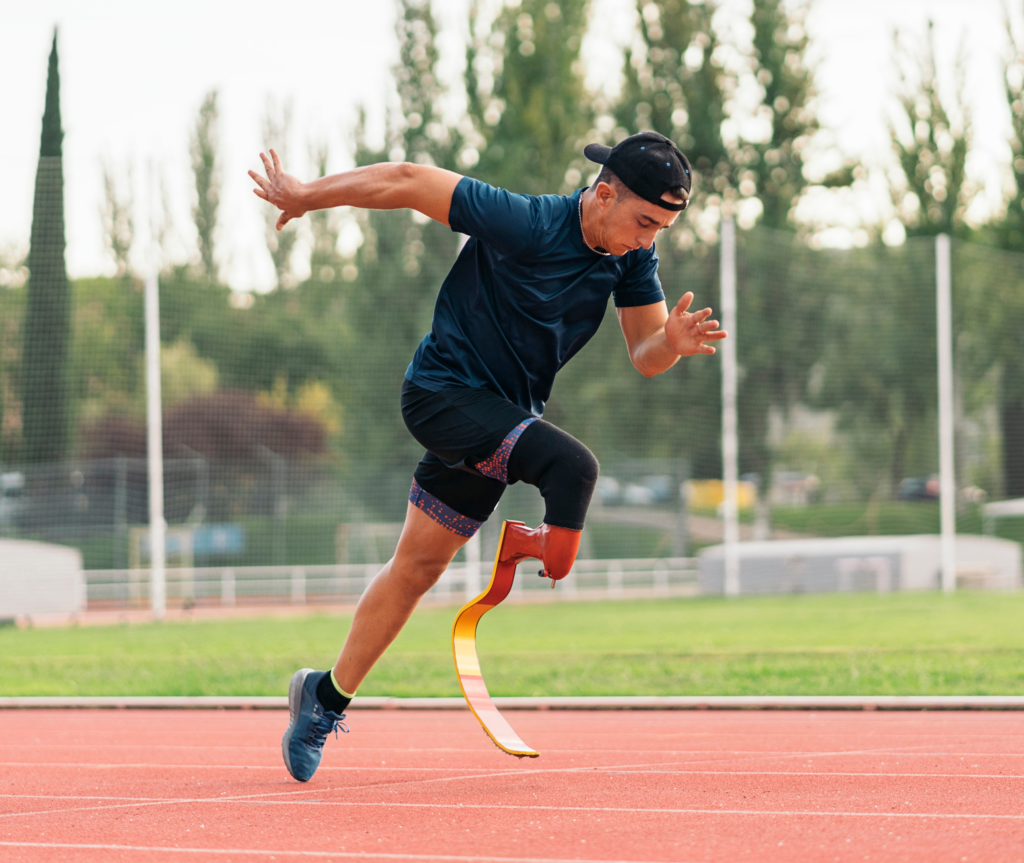 A man with a prosthetic leg runs on an athletics track. He is wearing a backwards hat, a black t-shirt, and shorts. The track stretches behind him, lined with green grass on either side.