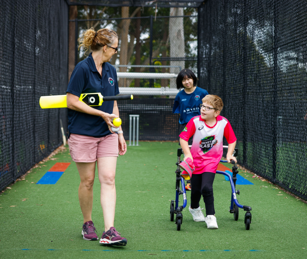 A woman walks with a kid wearing a Cricket Blash pink shirt. She holds a cricket bat under her arm and cricket balls in her right hand. The kid, using a walker, faces the camera and is engaged in conversation with the woman, who is the coach. The coach talks to the child while walking, with her back to the camera. Behind the child stands another person observing the conversation. They are on a cricket pitch with wickets in the background.