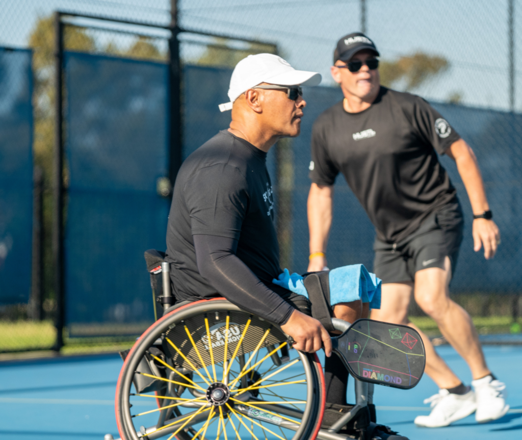A person in a wheelchair is holding a pickleball racket. He is wearing a white cap and sunglasses, looking away from the camera with a focused expression. In the background, another person is running backwards. This person is wearing a black shirt, black shorts, sunglasses, and a black cap.