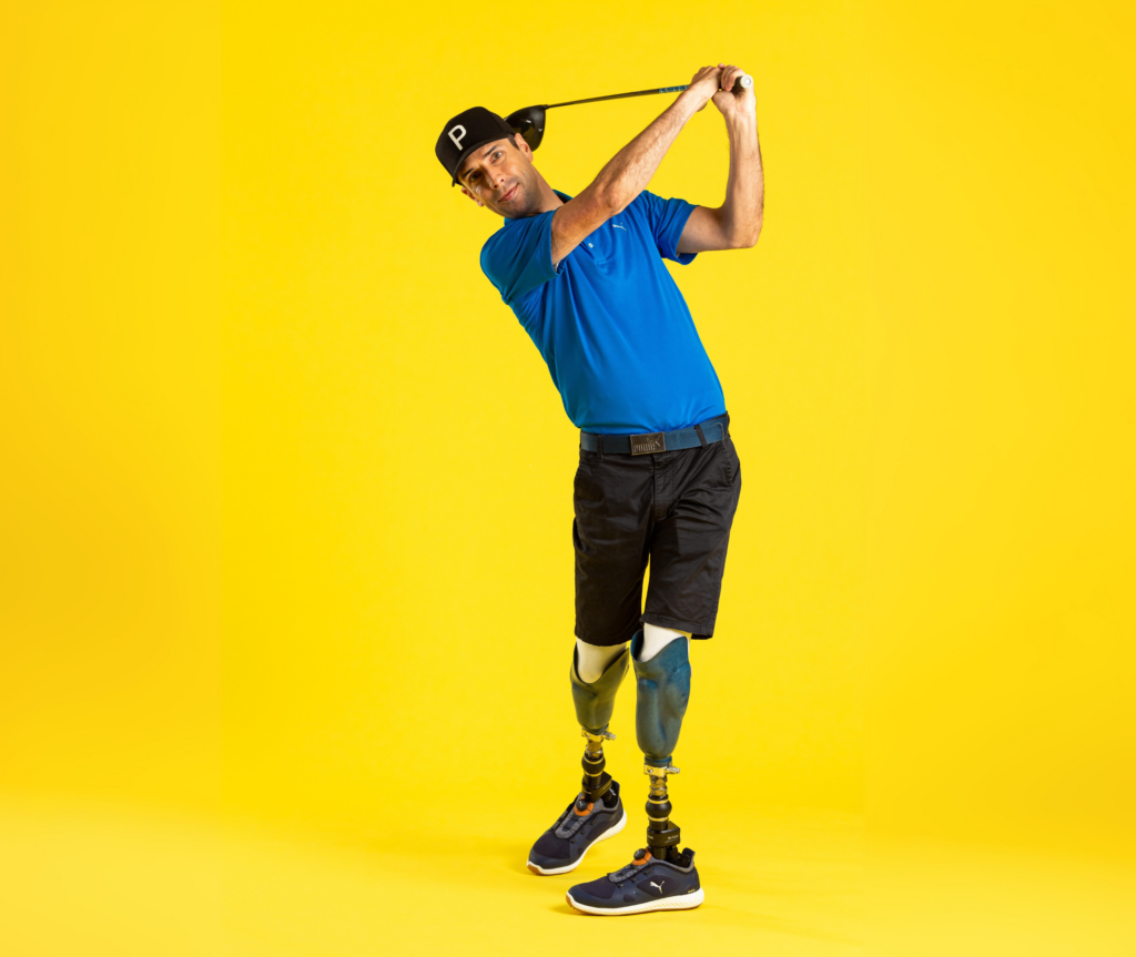 The image shows a Caucasian male with prosthetic legs. He is wearing a black cap, black shorts, and a blue shirt. He is in a pose indicating he has just hit a golf ball off the tee. The man is looking towards the camera with the pose held.