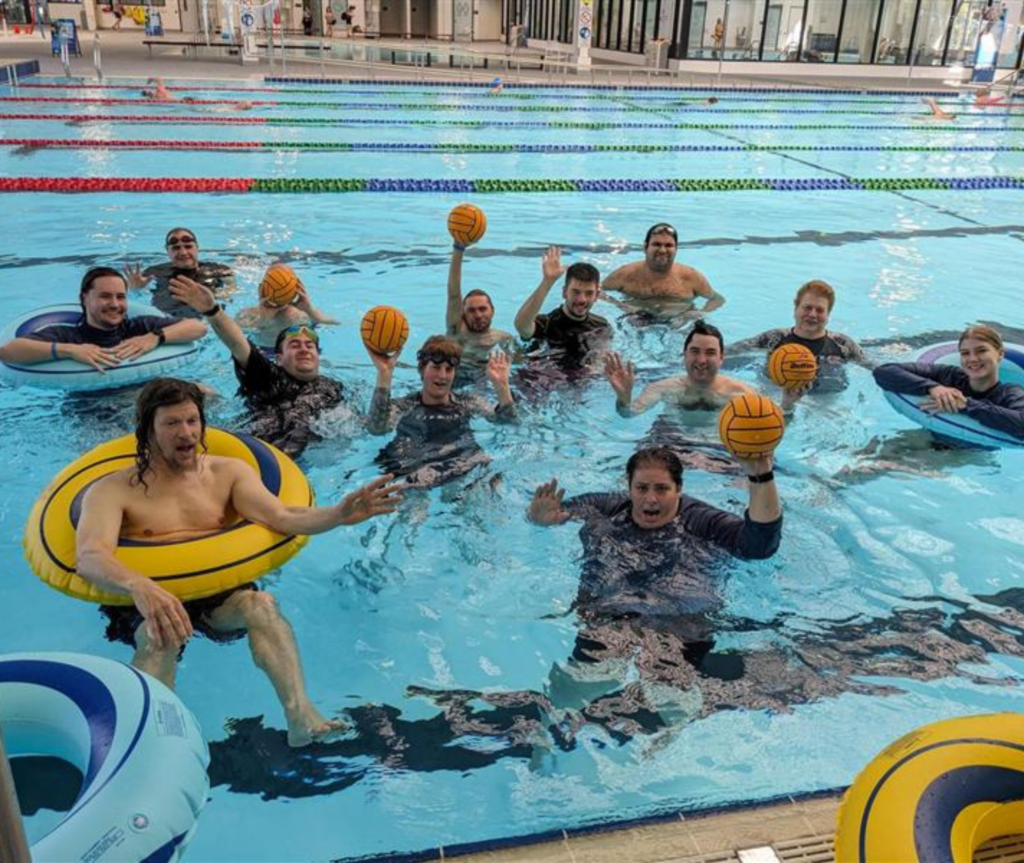 A diverse group of people in a pool, some with disabilities, holding a water polo ball and smiling together.