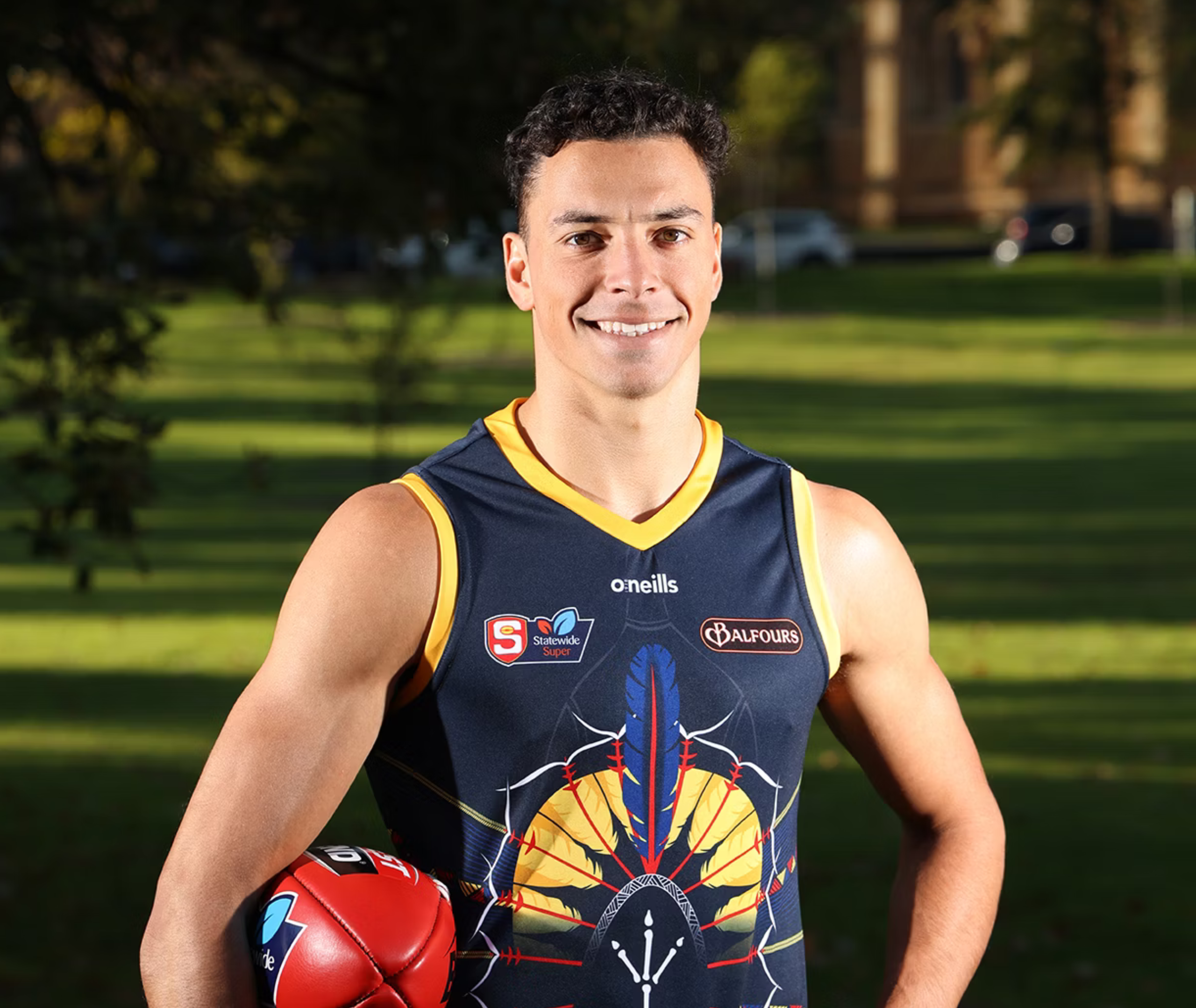 Close up image of Benjamin. He is wearing adelaide crows top and holding a footy under his arm.