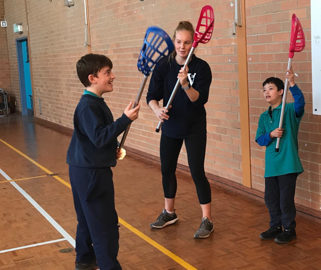 In the image, three people are shown at a lacrosse training session. In the center, the coach stands, demonstrating technique to one of the students who is holding a lacrosse bat. The coach is actively instructing the student while showing them proper form. On either side of the coach, two students are mimicking the coach's actions, also holding lacrosse bats as they practice the demonstrated technique.