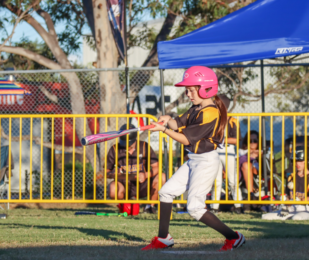 This image features a young girl wearing a brown shirt and white pants on a baseball field. She is wearing a pink helmet and holding a pink baseball bat, captured in an action shot where she has swung the bat and is about to hit the baseball. In the background, other teammates and a coach can be seen, blurred at a distance.
