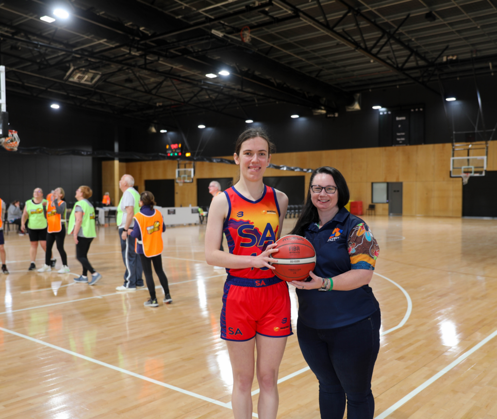 Sport4All's Inclusion Coach Kelly with a Basketball SA athlete, both holding a basketball and smiling at the camera. In the background, some people are playing basketball, but they are blurred.