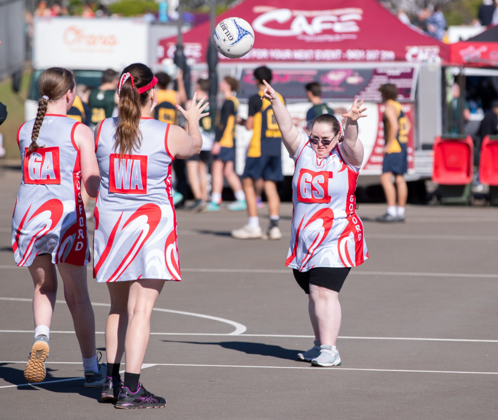 The image shows a woman with dark hair wearing a white netball kit and sunglasses. She is throwing the ball to two other girls from the same team who are standing opposite her with their backs to the camera. They are all wearing the same team kits. They are on a netball court with other players in the blurred background.