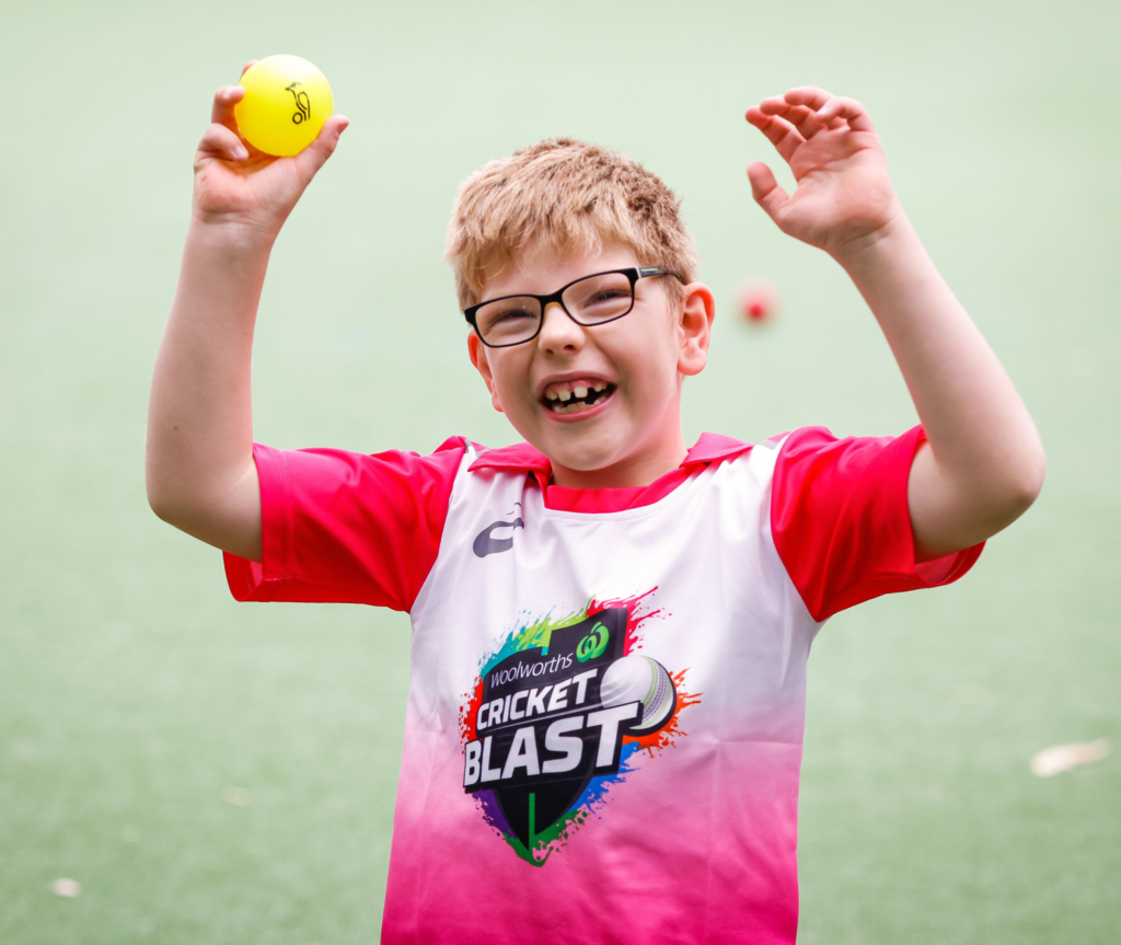 In this imagxe, a young boy is wearing a pink Cricket Blast jersey. He is wearing glasses and is smiling while looking at the camera. He has both his hands in the air with a cricket ball in his right hand. He is on a cricket field, which is visible in the background.