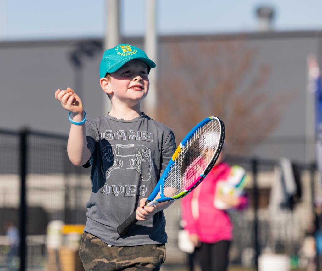 A young boy holding a tennis racket in his hand. He is wearing a black cap and grey shorts. He is smiling and is on a tennis court.