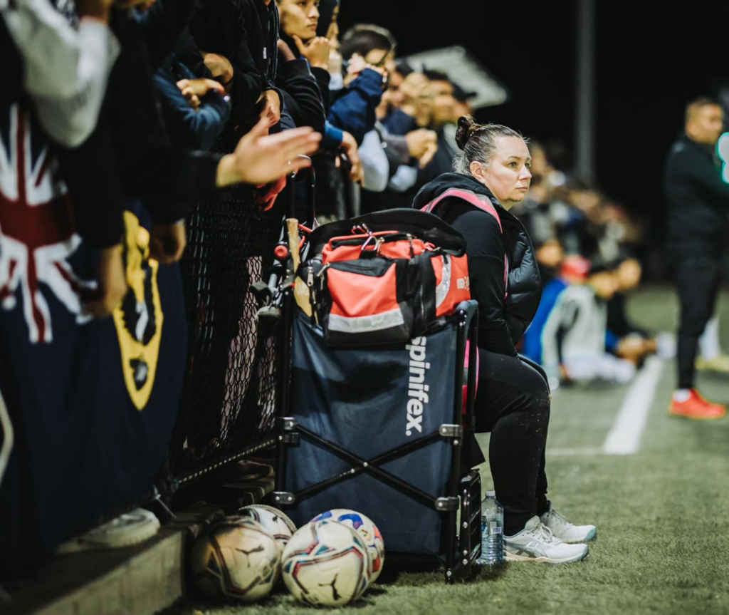 An image of Kelly seated by the soccer pitch, next to a fence. She is wearing a black tracksuit and looking at the game. Beside her are some footballs, and behind the fence, people are watching the game.