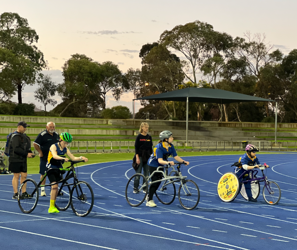 An image showing para cyclists on a racing track, with their bicycles lined up on the track. Several coaches stand behind them, observing and offering support. The cyclists wear helmets and racing gear, while the coaches are dressed in casual sports attire. The background features elements of a standard racing track, including lanes and markings.