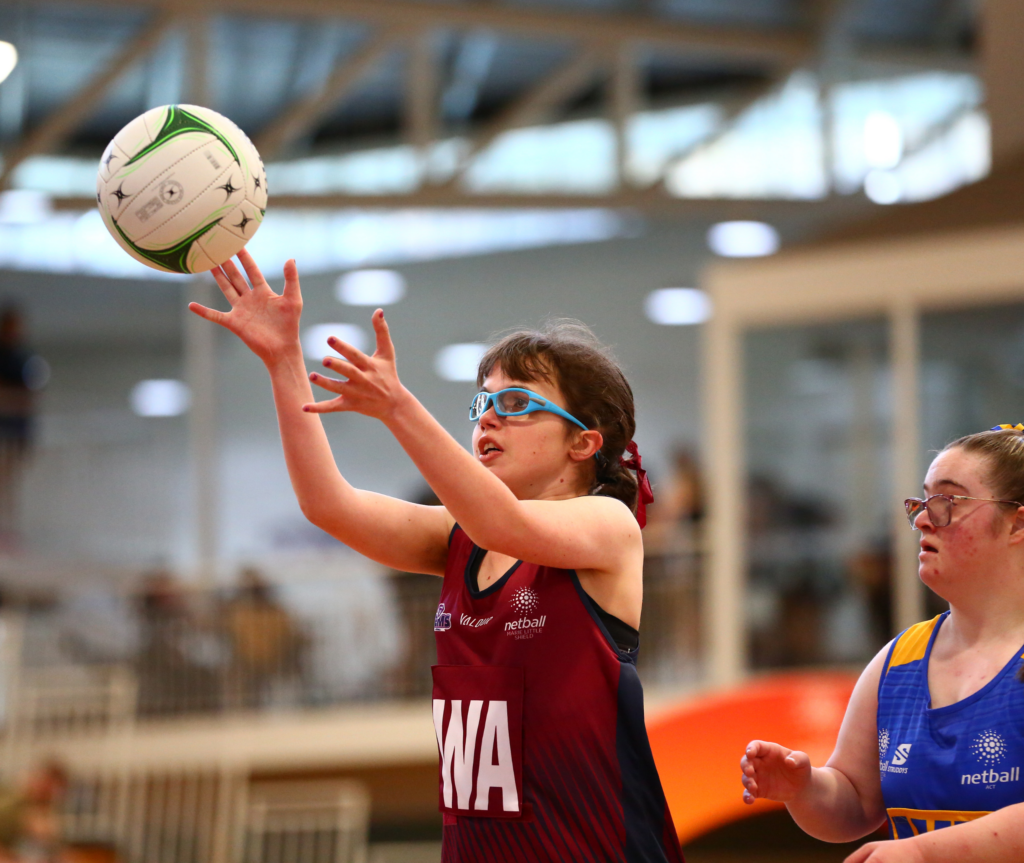 Two girls playing netball on a court, both wearing protective glasses, one in red and the other in blue.