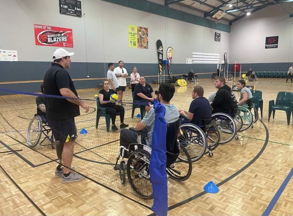 A group of participants in wheelchairs on a volleyball court, preparing to play wheelchair volleyball. A coach stands beside them, engaging in a discussion and offering guidance.