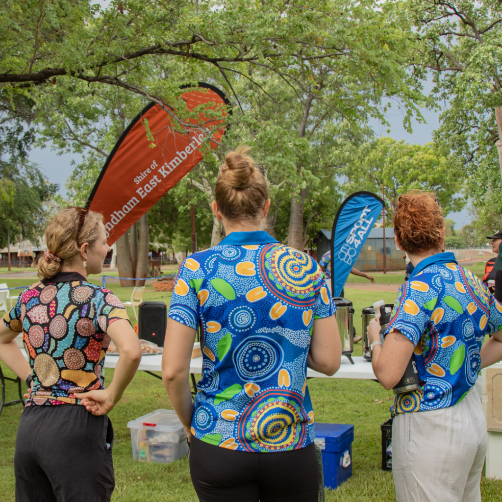 Three SWEK staff members stand with their backs to the camera, wearing polo shirts. The SWEK and Sport4All banners are visible in the background.