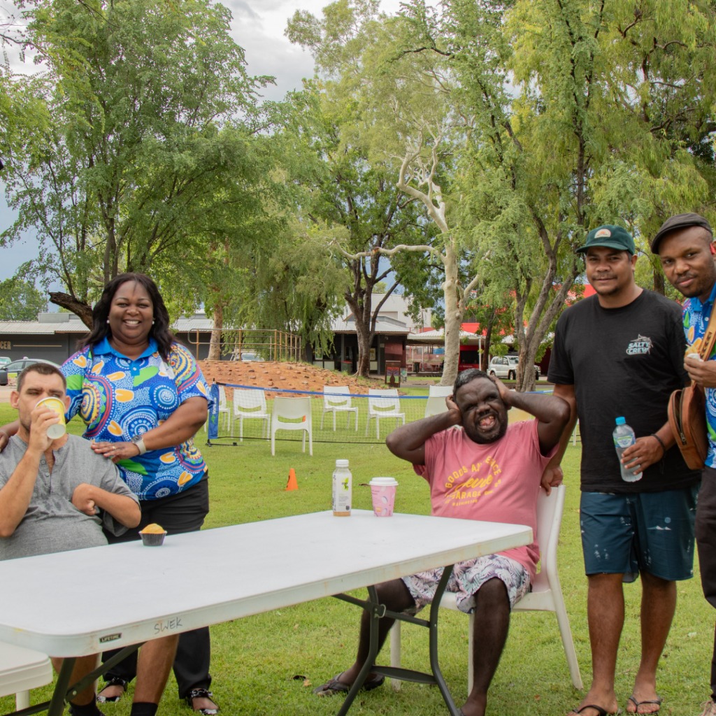 Participants sit on chairs next to a table, surrounded by SWEK council staff wearing colourful First Nations-themed polo shirts. The group is posing for the camera.