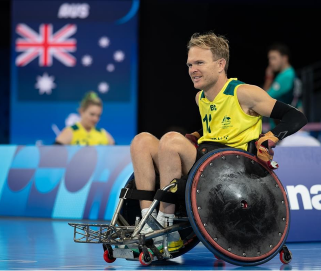 Beau Vernon, Sport4All Inclusion Coach, in a wheelchair at a sporting event wearing the Australian kit. He is looking away from the camera with his hands on the wheelchair. A faded Australian flag is in the background.