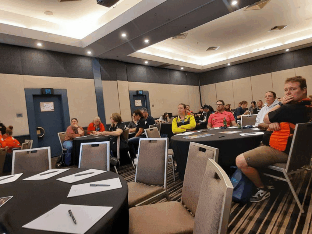 A group of people sit at round tables in a conference room, attentively listening to a presentation.