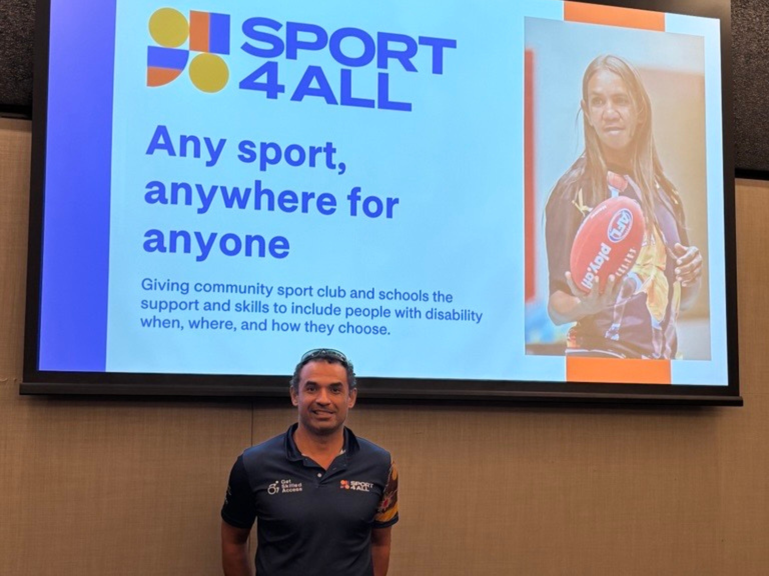 Matthais, Sport4All Inclusion Coach, in a Sport4All polo shirt stands in front of a presentation slide that reads “Any sport, anywhere for anyone” with the Sport4All logo and a photo of a woman holding a football.