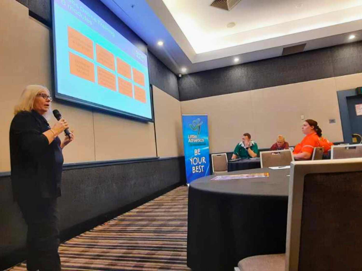 Kim, Sport4All State Manager, presents at a workshop, speaking into a microphone in front of a screen showing inclusion training content.