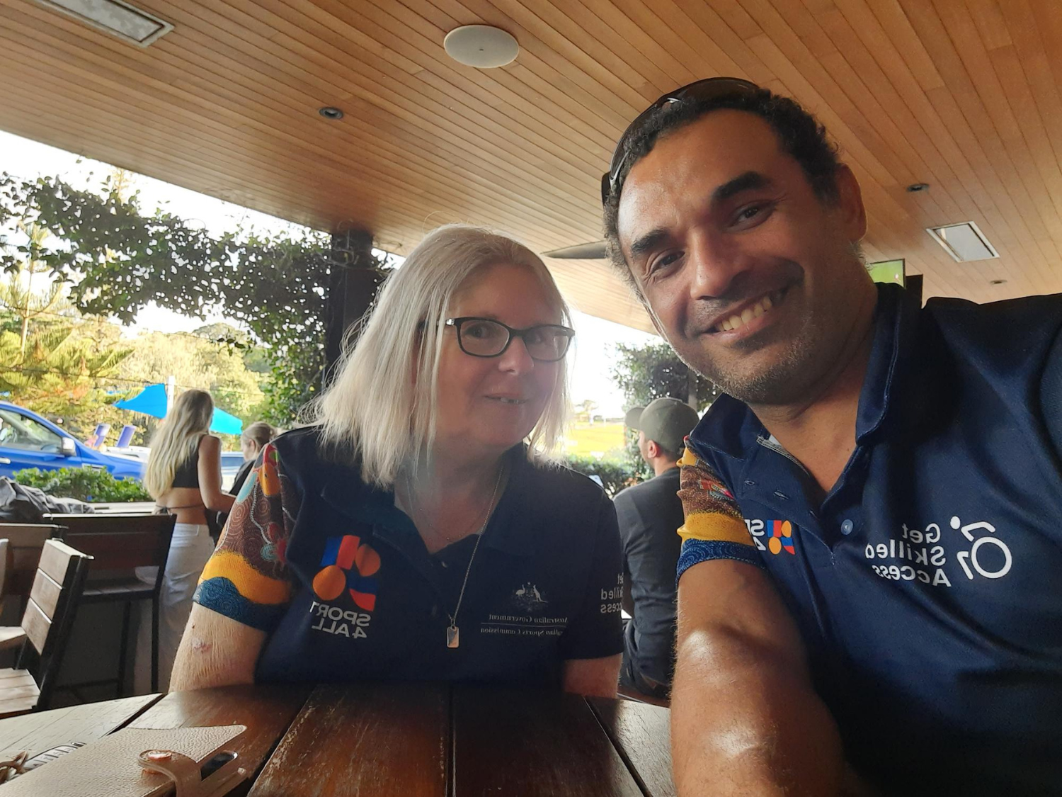 Kim and Matthais wearing Sport4All shirts smile for a selfie at an outdoor café.