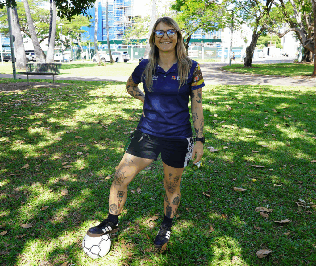 Sport4All Inclusion Coach, Alex wearing Sport4All polo in an outdoor setting looking at the camera and smiling, She has a football under her feet.