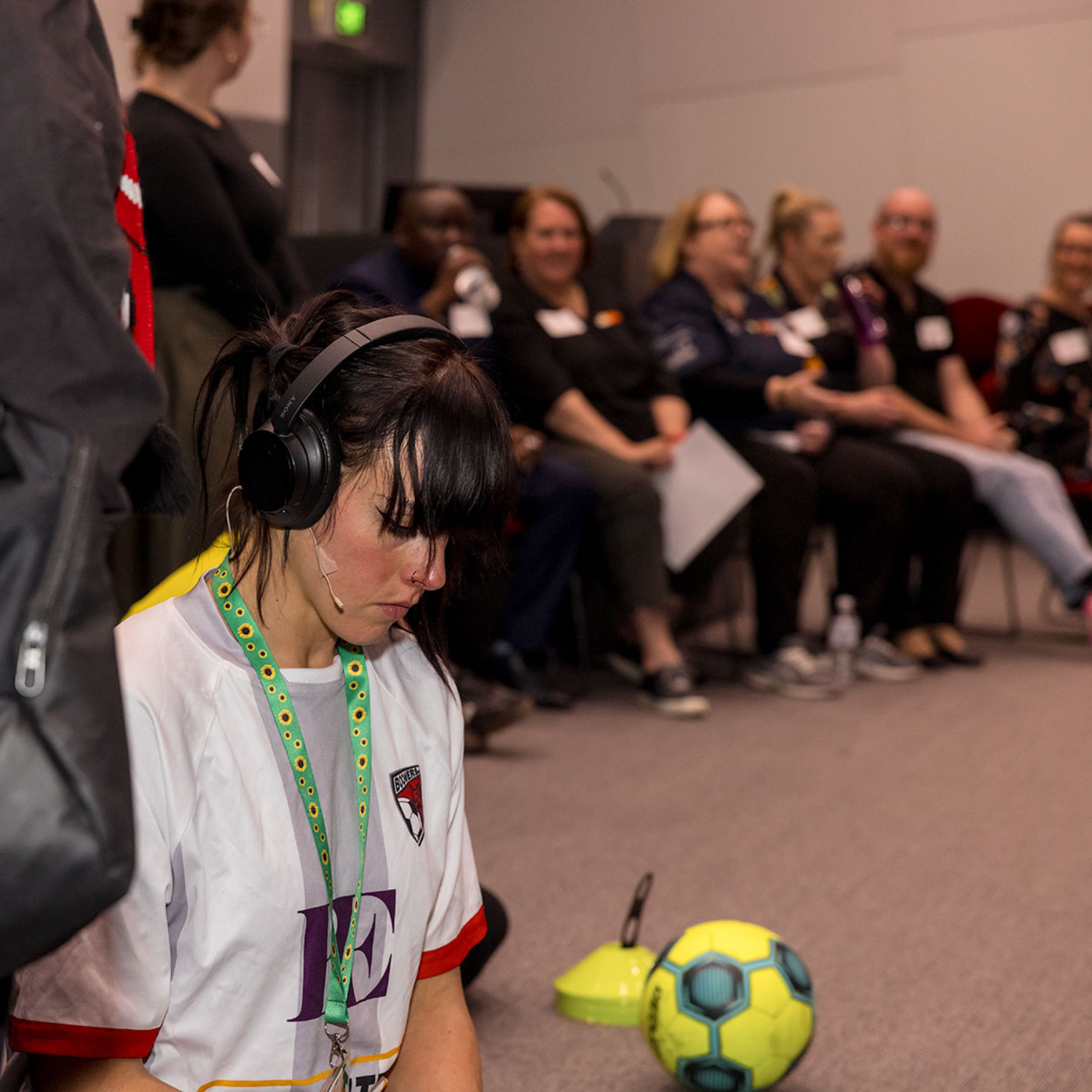 Young woman wearing sunflower lanyard, headphones and a sports jersey sits on the floor near a football, with a group of people seated in the background watching.