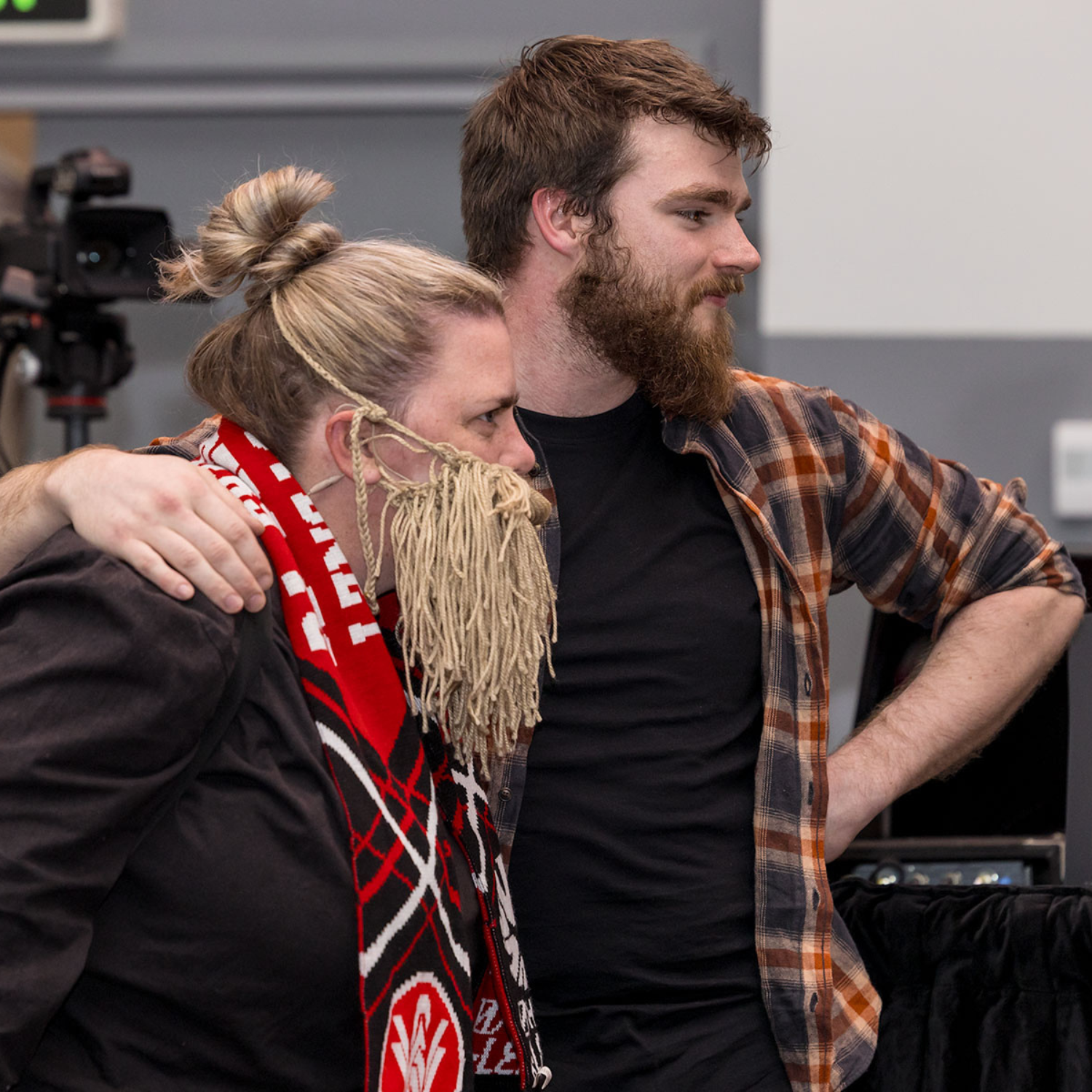 Two people standing close together, one with their arm around the other. One person wears a fake yarn beard and a red and black scarf, while the other has a real beard and wears a checked shirt.