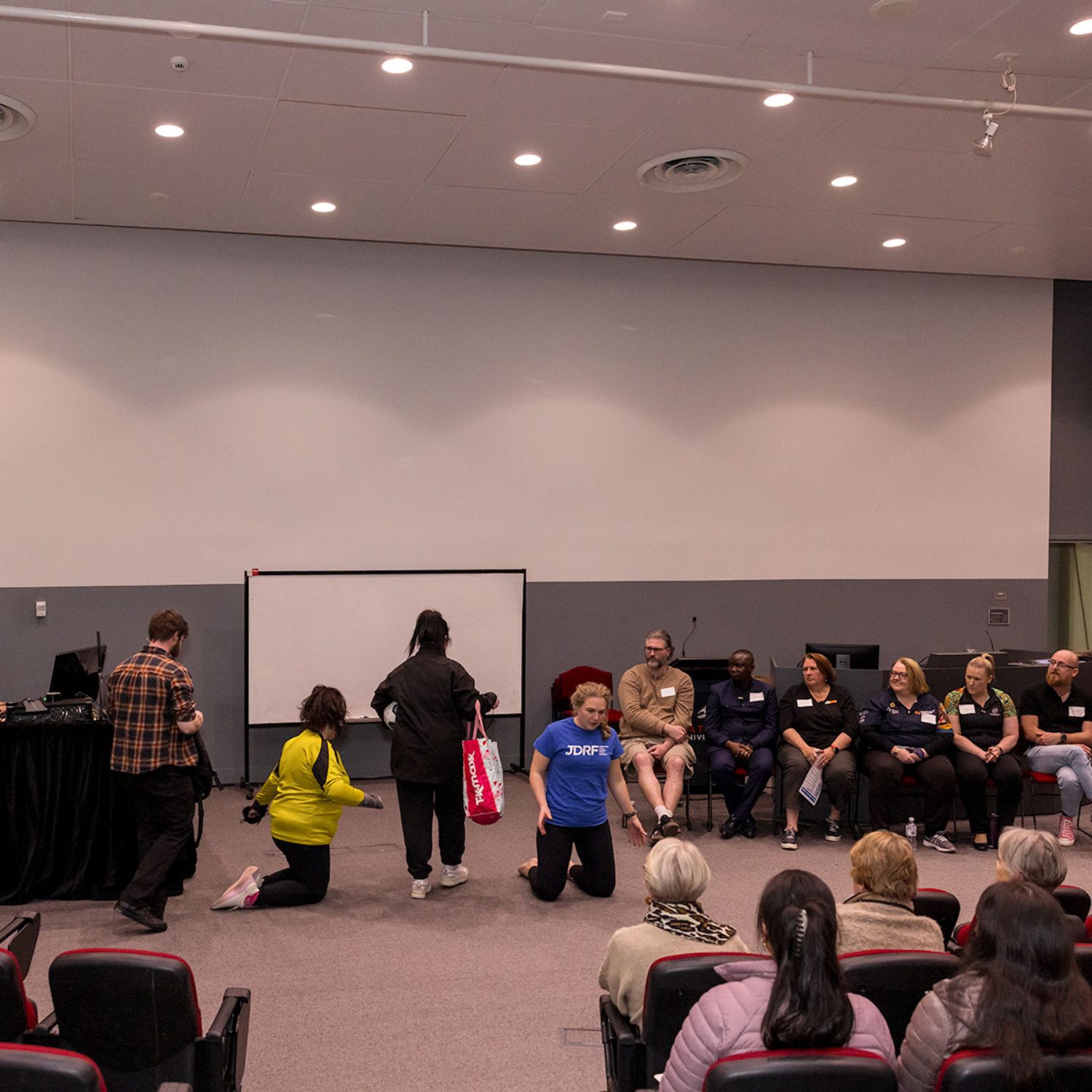 Wide shot of a staged activity in a lecture hall. Several people kneel or stand at the front while an audience watches. A row of participants sit along the wall observing.
