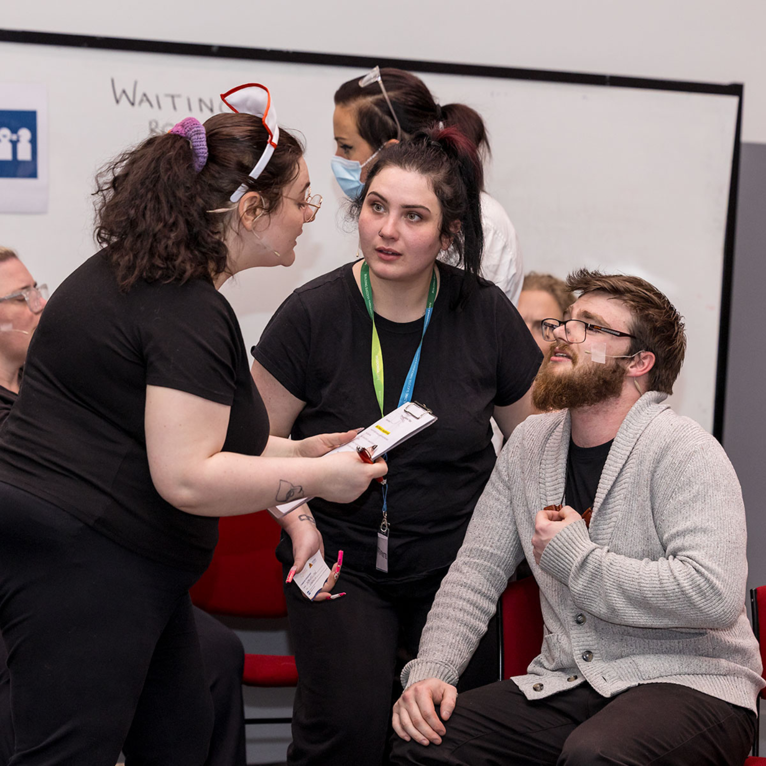 Three people in black shirts engaging in a role-play activity. One person holds a clipboard while speaking to another, who is seated with a bandage on their face, as a third listens attentively.