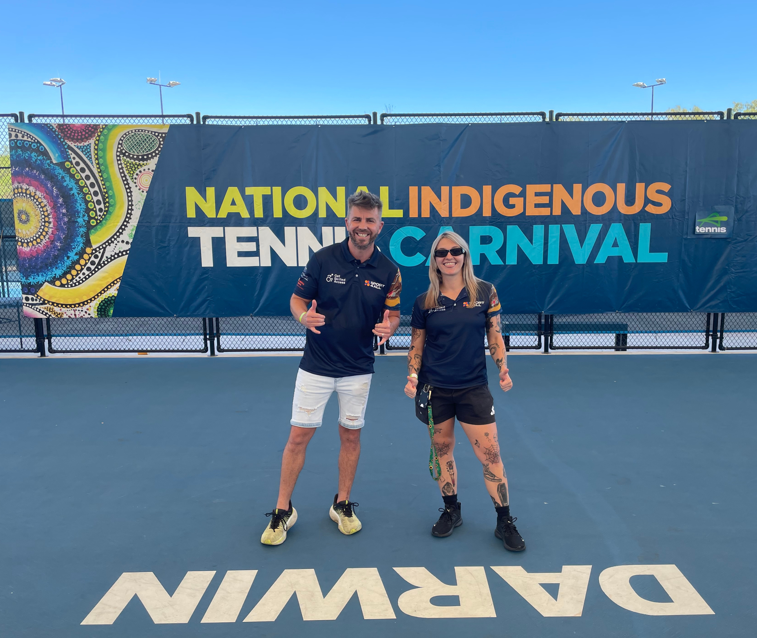 Sport4All Inclusion coaches, Alex and John are wearing Sport4All shirts standing in front of a large banner that reads “National Indigenous Tennis Carnival” on a court marked with the word “Darwin.”