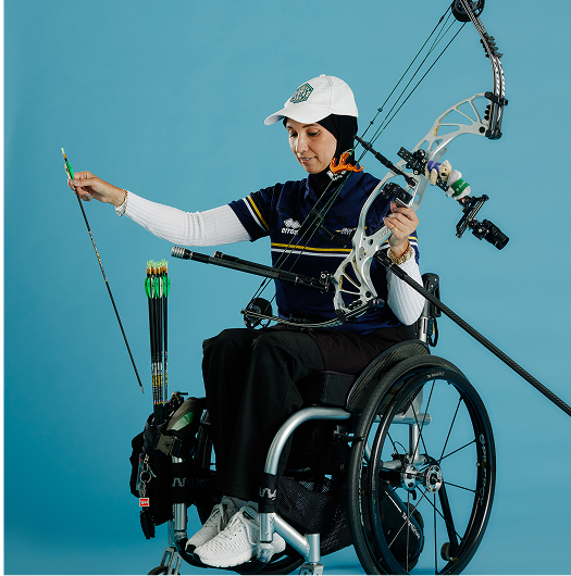 An archer on wheelchair wearing blue kit and a white hat is drawing an arrow and has the bow in one hand.
