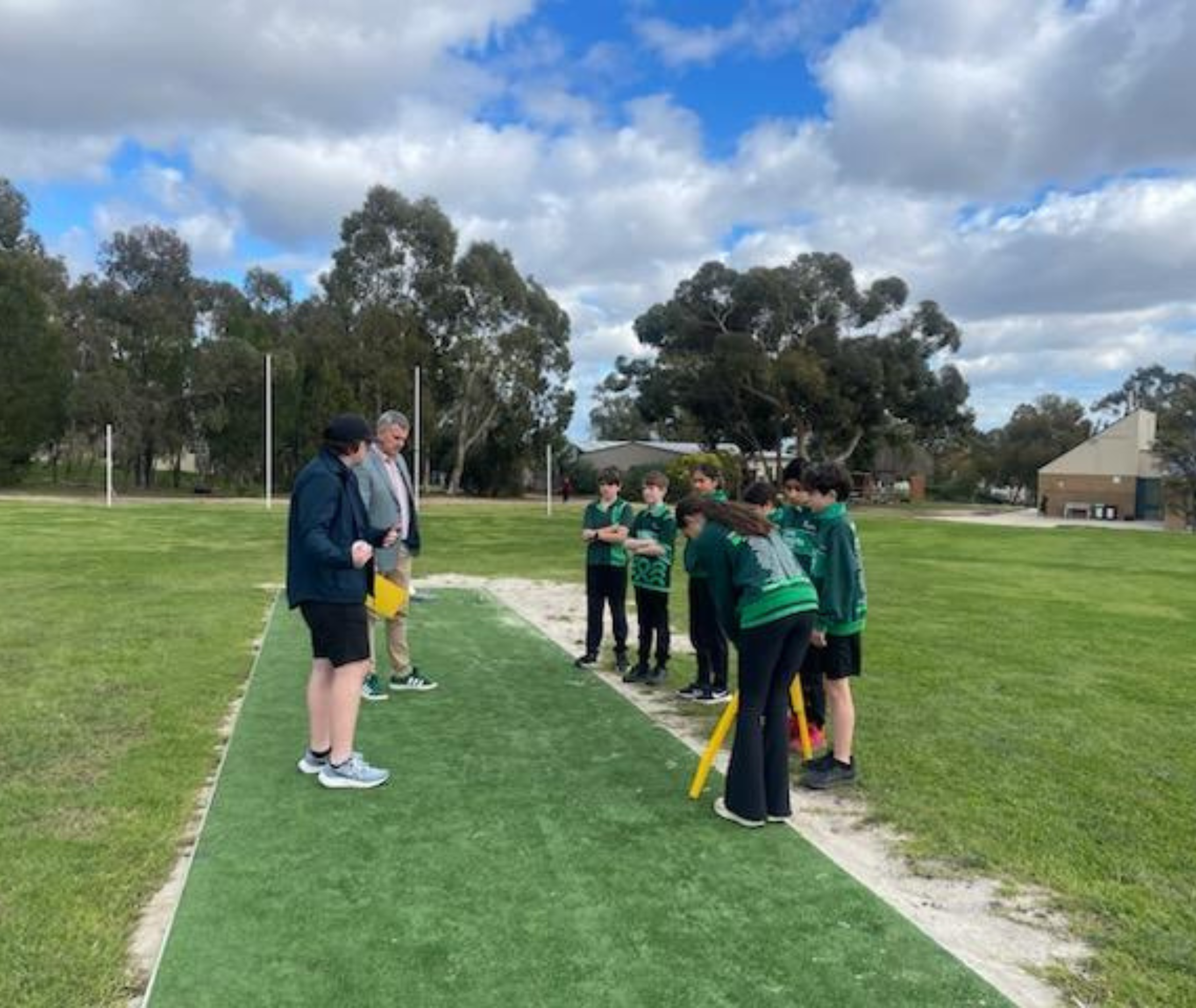 Students playing cricket on an outdoor pitch, wearing yellow simulation glasses, with other students participating in activities in the background.