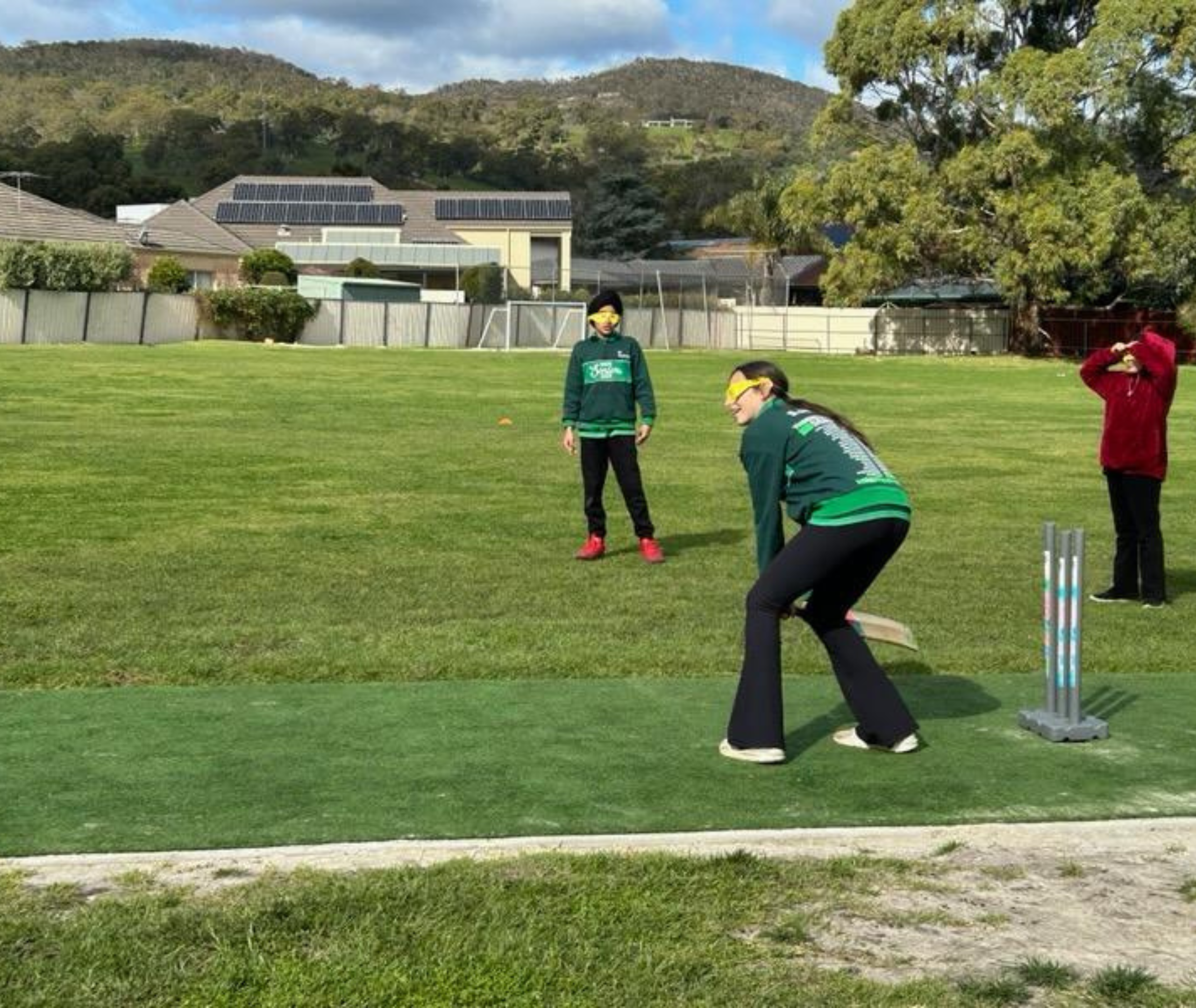 Student in green sports uniform batting while wearing yellow simulation glasses, with other students and an adult in red jacket standing in the background.