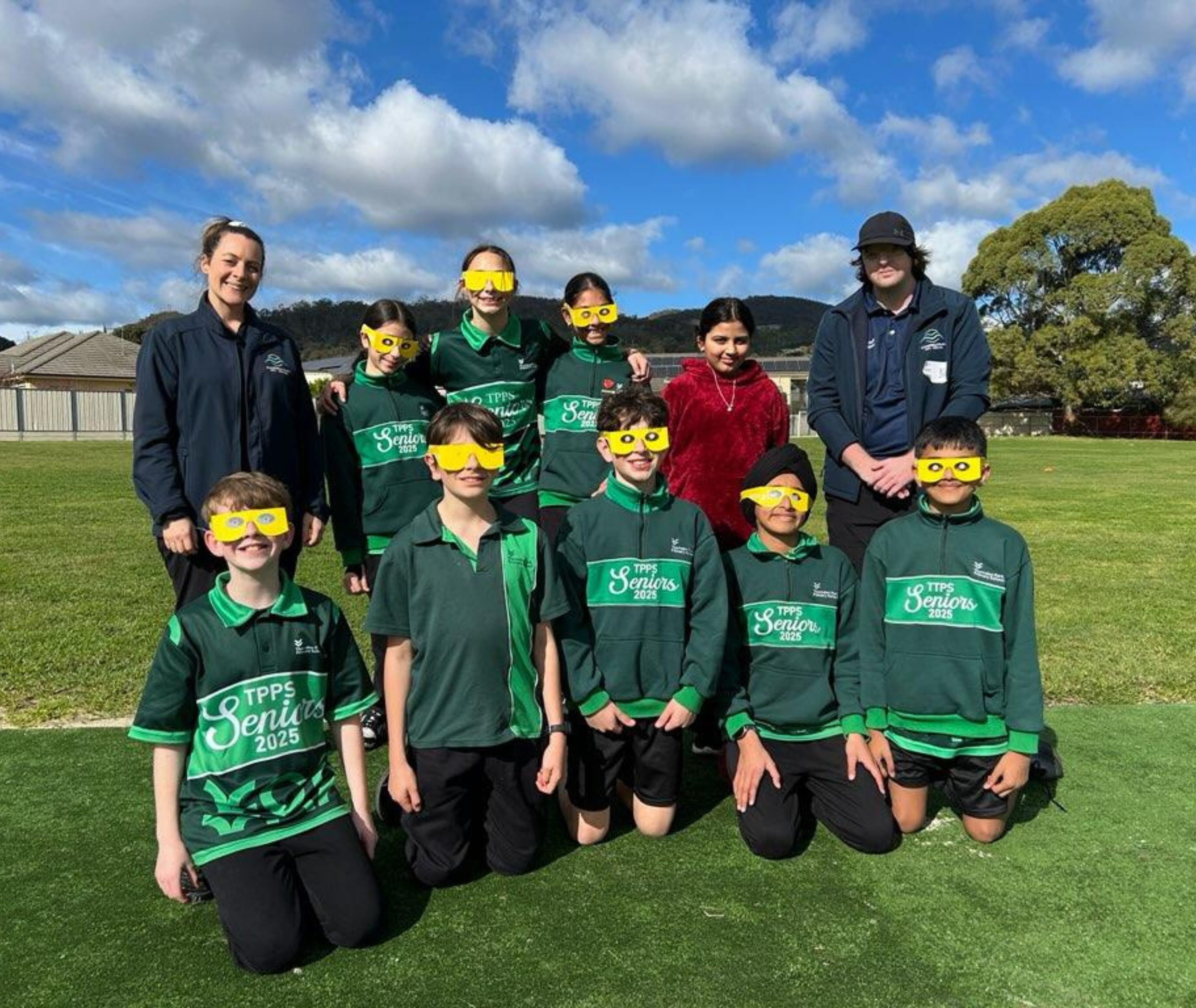 Group of students in green uniforms and two adults posing together on a grass field, all students wearing yellow simulation glasses.