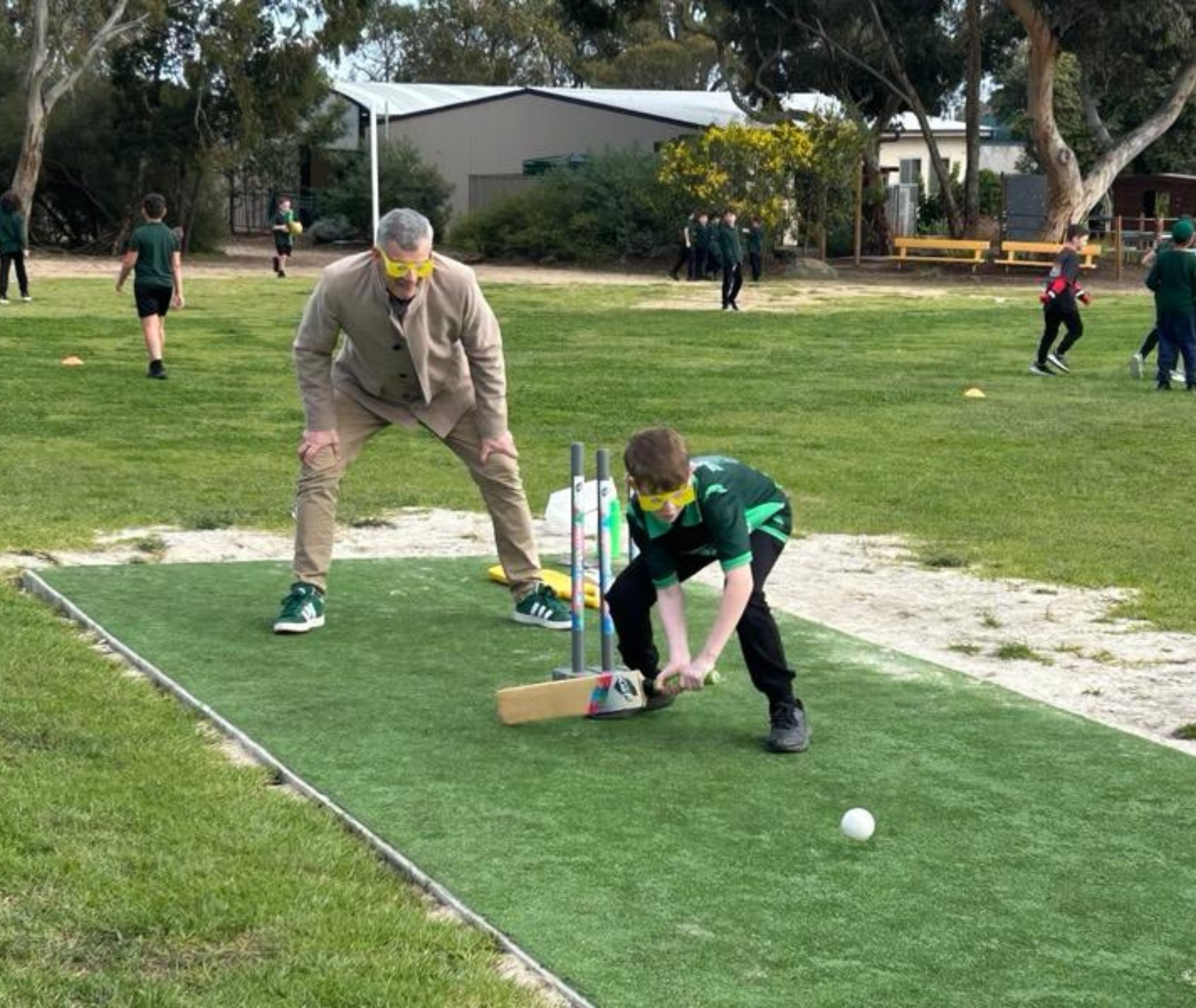 Student in green sports uniform batting while an adult in beige suit stands behind as wicketkeeper, both wearing yellow simulation glasses.