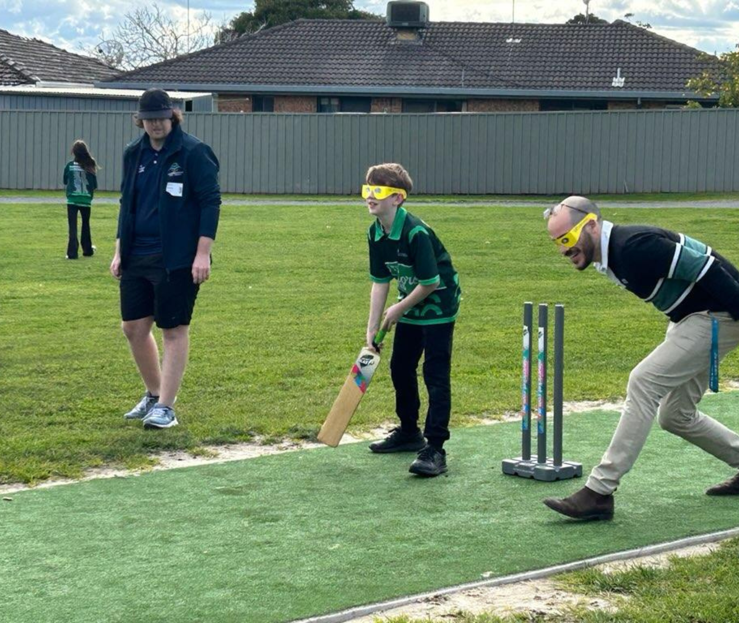 Student preparing to bat with yellow simulation glasses while an adult bends to field and another adult stands nearby observing.