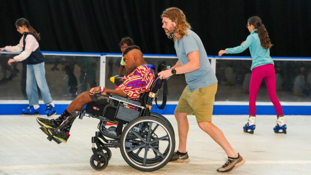 A person in a wheelchair being assisted by another person pushing from behind on an ice skating rink in Alice Springs. In the background, others are also enjoying ice skating.