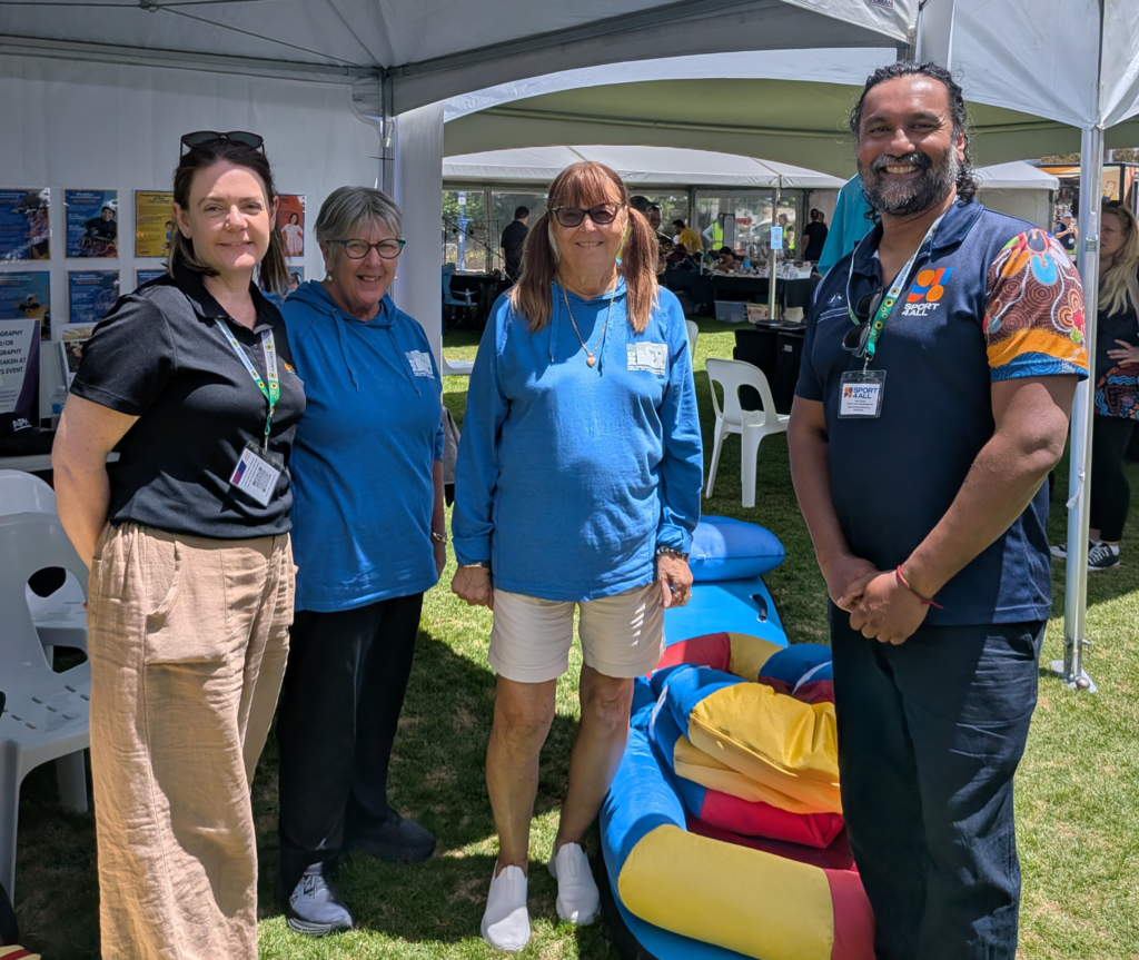Four people stand together under a marquee at a community event, with Sport4All signage and colourful soft play equipment visible behind them.
