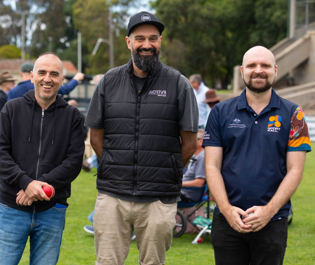 Three men stand side by side on a grass oval at a community cricket event - to the right of the image is Nathan Walsh in Sport4All top, smiling toward the camera, with other participants and spectators in the background.