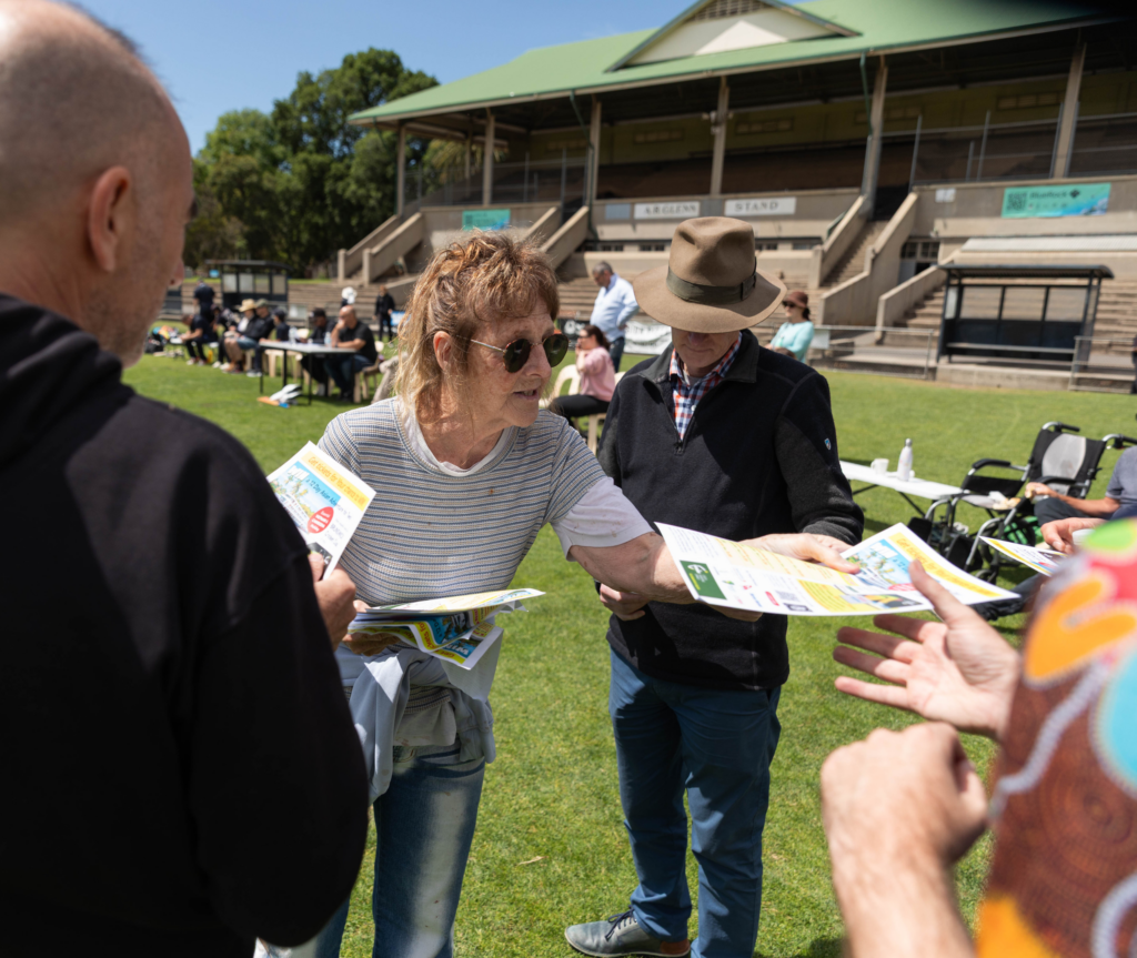 People stand together on a grass oval handing out flyers and talking with attendees, with stadium seating and event tables behind them.