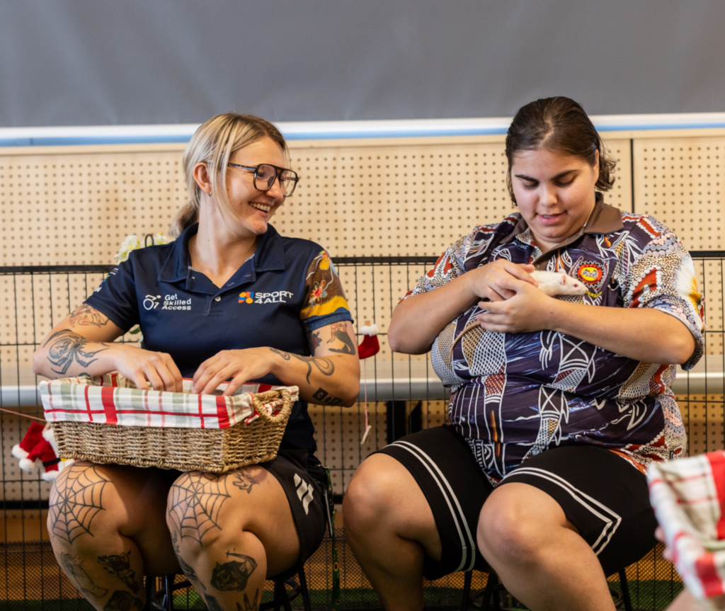 Two people sit side by side indoors; Sport4All Inclusion Coach, Alex, wears a Sport4All polo and smiles while the other gently holds a small animal, with baskets and seating visible around them.