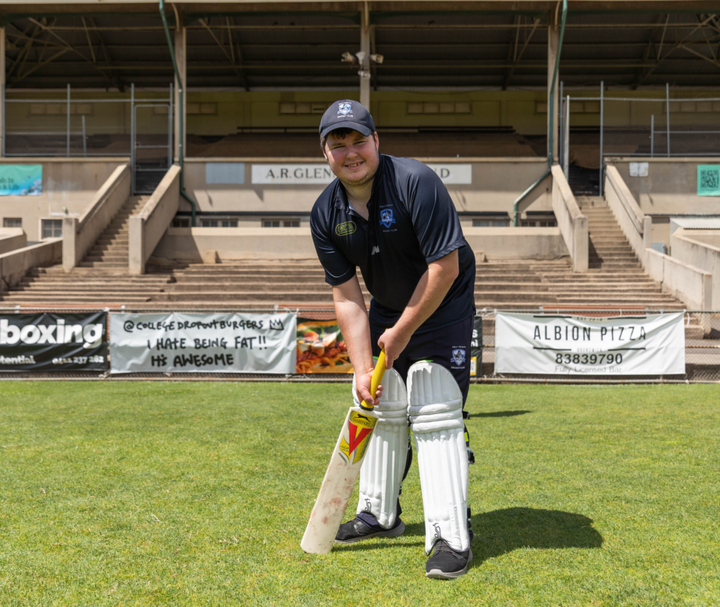A cricket player stands at the crease holding a bat, wearing leg pads and a helmet, with stadium seating visible behind them.