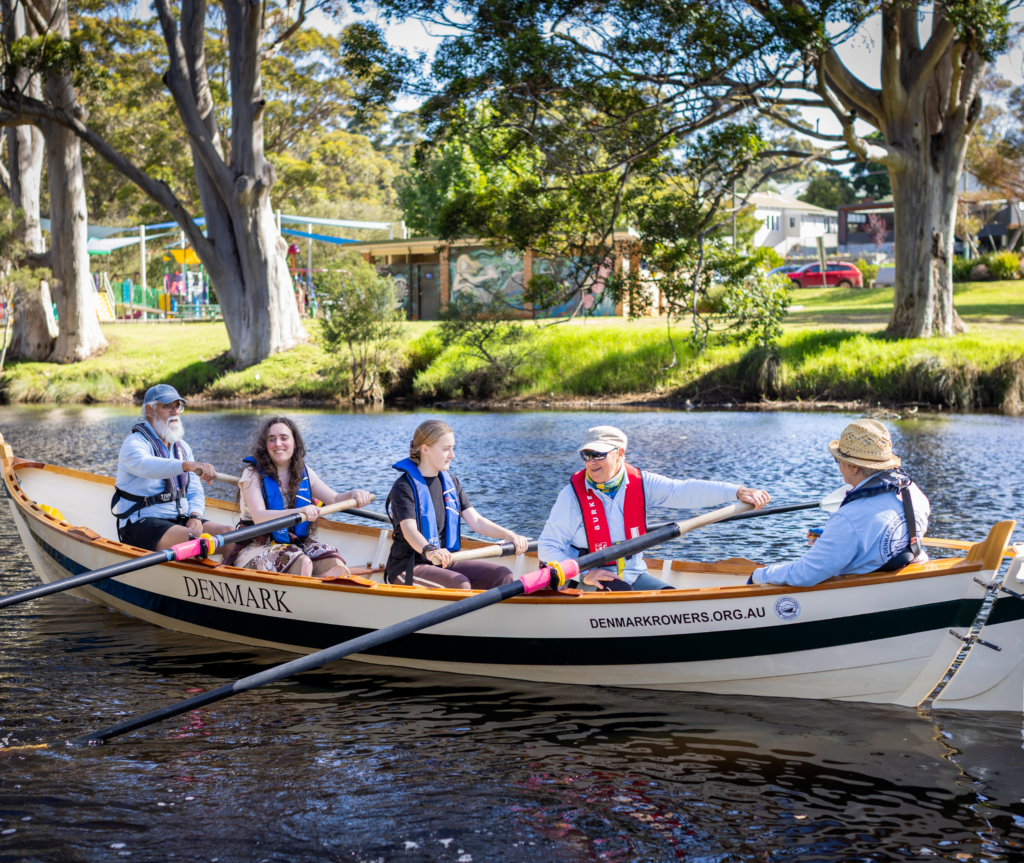 A mixed group of people row together in a boat on a calm river, wearing life jackets, with trees and parkland visible along the riverbank.