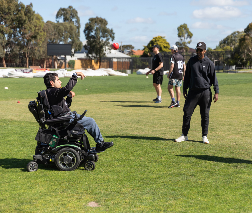 A participant using a powered wheelchair throws a cricket ball across a grass oval during a modified cricket activity, with others watching nearby.