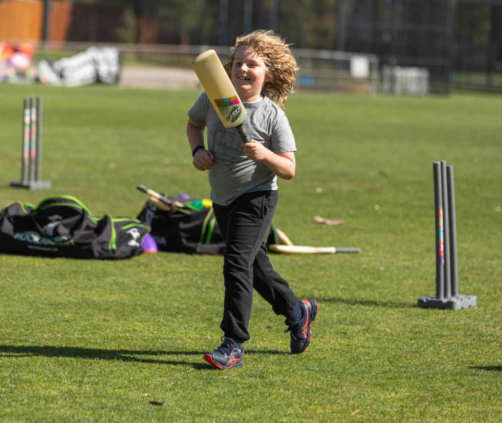 A young child runs across a grass oval holding a cricket bat, smiling, with training equipment and bags visible in the background.