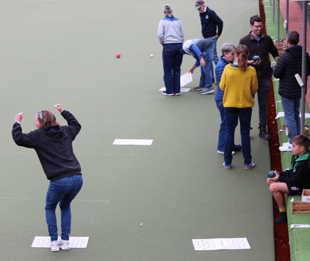 A group of people playing lawn bowls on an outdoor green. One person in the foreground celebrates with raised arms while others chat and take turns bowling along the edge.