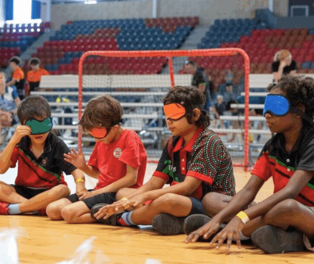 Young students sit on a court wearing blindfolds and listening for the sound of the goalball. A goal net and spectators are visible in the background.