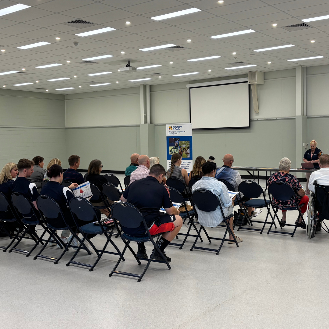A group of adults sit on chairs in a hall listening to a Sport4All presentation.